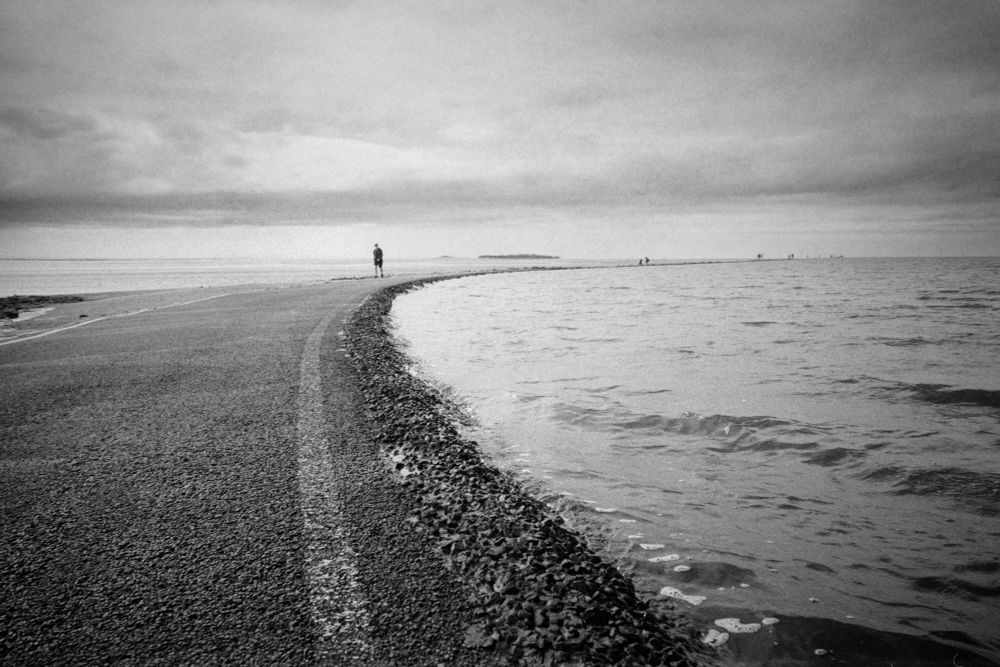 Black and white photo of a walkway between two bodies of water, it is curving away to the right, disappearing into the distance, where a lone figure is walking towards the camera. 
