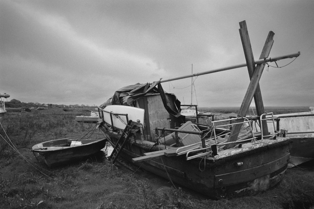 B&W photo of an old patrol boat, layed up on a marsh, the boat has been used as a place to live, a refuse bin in the rear deck, and a mast used as a place to dry clothes.  
