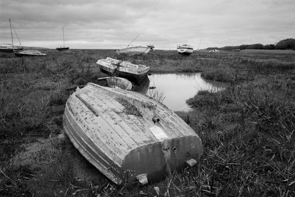 B&W photo of many boats varying in size, that have been abandoned on a salt marsh close to a river inlet. A pool of water reflects the dull grey autumnal sky.   