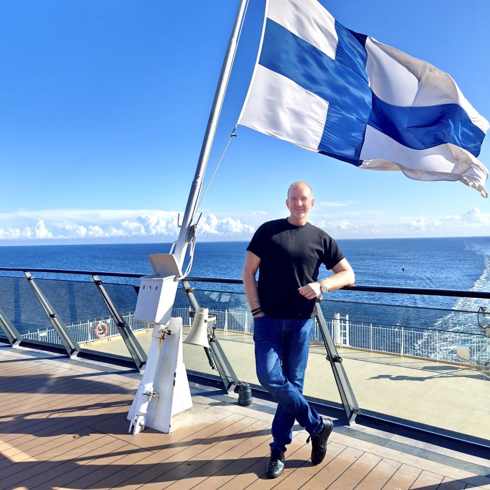 Jan Holmberg at the cruise ship on front of the blue sky, white clouds, sea and Finnish flag.