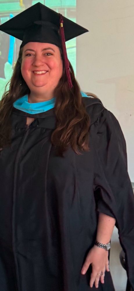 A zoomed in picture shows MsGillELA still in a cap and gown, this time with her light blue education hood over her shoulders and in front of her neck. You can see the projected graduation ceremony in part of the background with no identifying info of other students. 