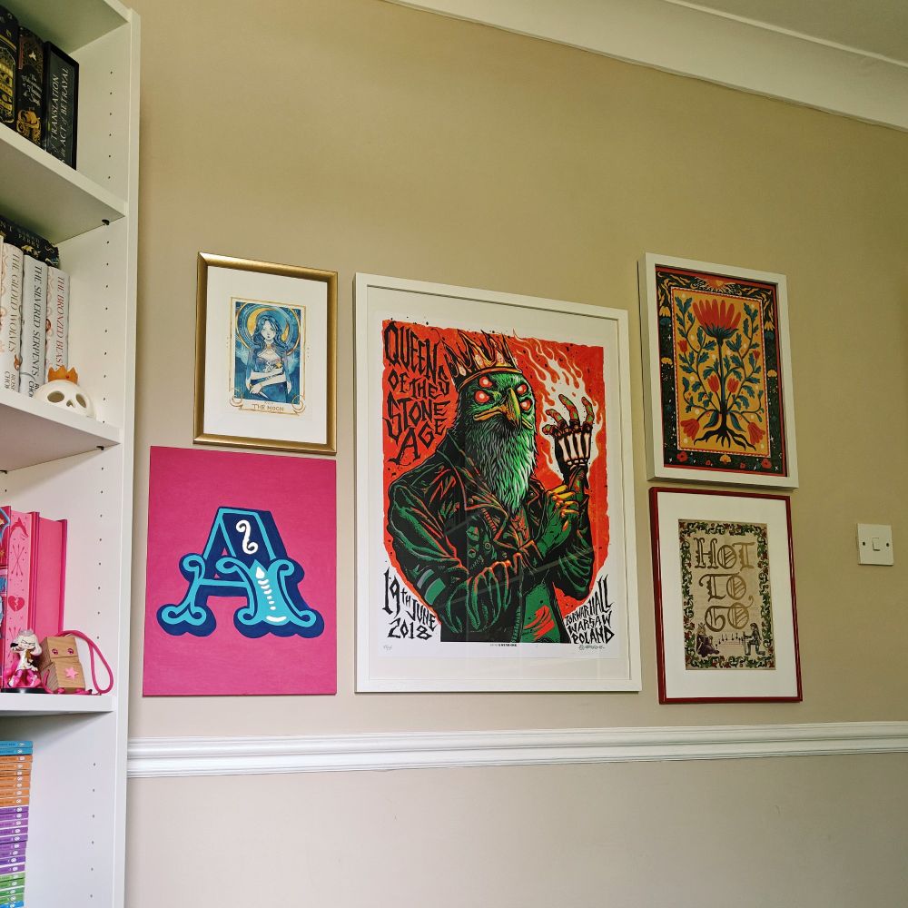 Five art pieces on the wall, top to bottom left to right: A. a small watercolour illustration of a Moon tarot card by Feefall B. a sign style large letter A with foil detailing against hot pink background done by Anna C. a large poster for a Queens of the Stone Age show in Warsaw, Poland from 19 June 2018 by Munk One D. an art naouveau-esque print with a flower illustration by Sonny Ross E. a medieval style illustration for Chappel Roan's "Hot to Go" by Artis Marginalia