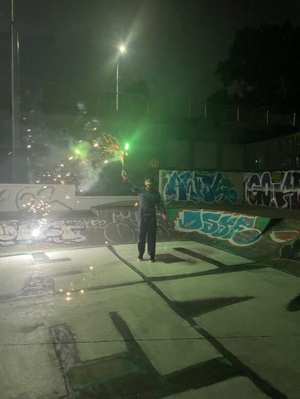 A man dressed in a Japanese school boy outfit for Halloween stands gleefully one hand pointed upwards grasping a firework that’s shooting eldritch green sparks. He stands in a skating bowl surrounded by artsy graffiti.