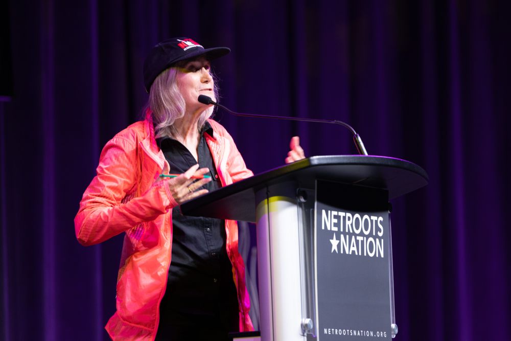 A woman in a baseball cap and red jacket, Amy Goodman stands speaking, at a podium with Netroots Nation on the front