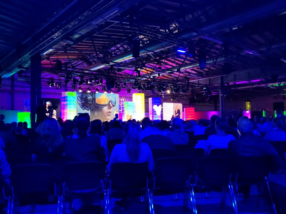 Ein Bild in einer Halle mit vielen Menschen und einer großen Bühne.