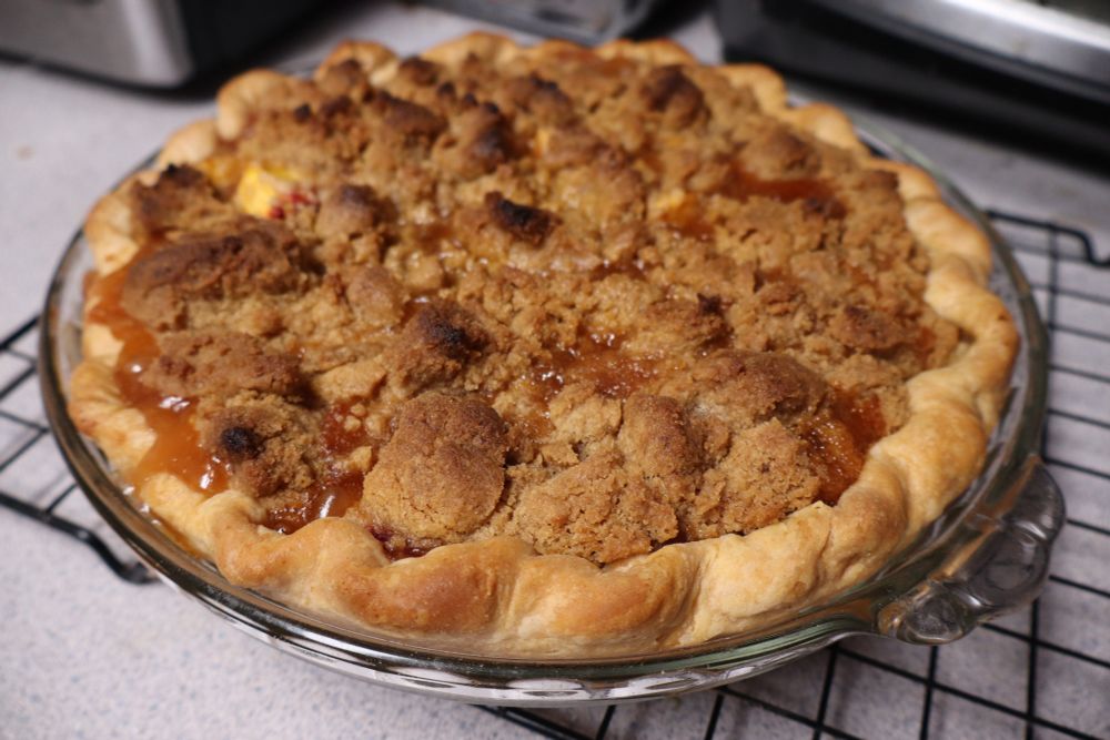 a whole pie on a wire rack. the bottom crust is fluted at the edges and the top is crisp, brown streusel. there's bits of orange goo peeking out from under it.