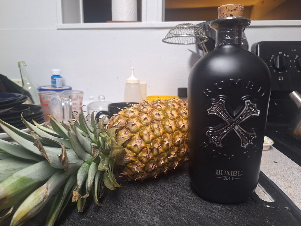 A pineapple beside a black bottle of Bumbu rum wit a cross symbol on it