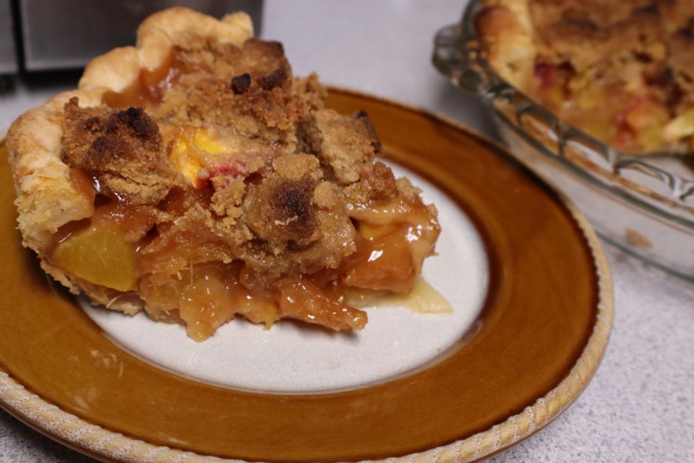 a slice of pie on a small plate. the bottom crust is fluted at the edges and the top is crisp, brown streusel. yellowy-orange chunks of peach and pineapple visible.
