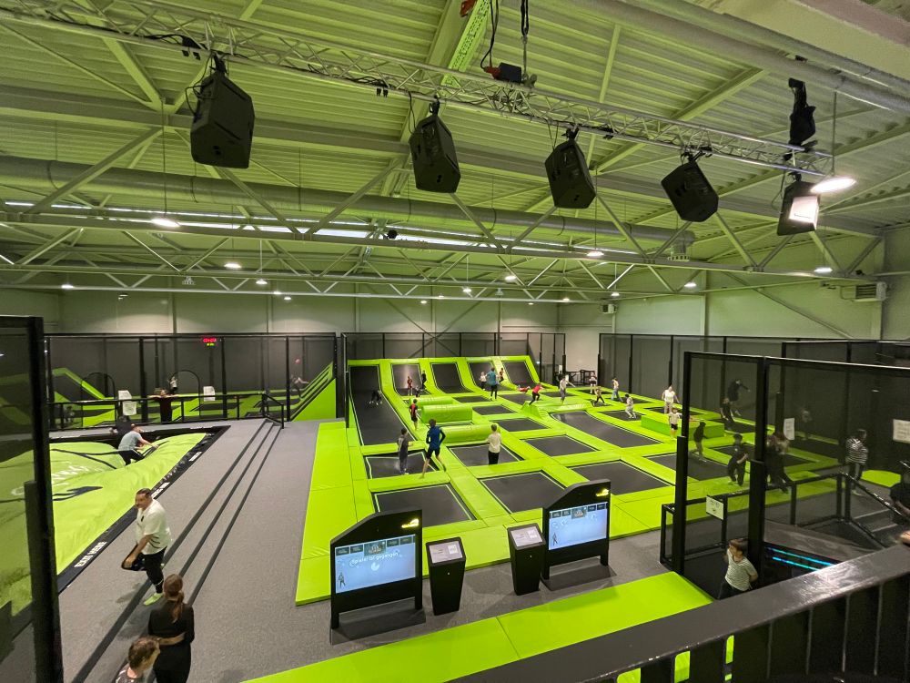 Eine Trampolinhalle. Unzählige Trampoline mit grüner polsternder Umrahmung. Viele Kinder und Jugendliche springen wild durcheinander. 
An der Decke hängen schwarze Scheinwerfer. 