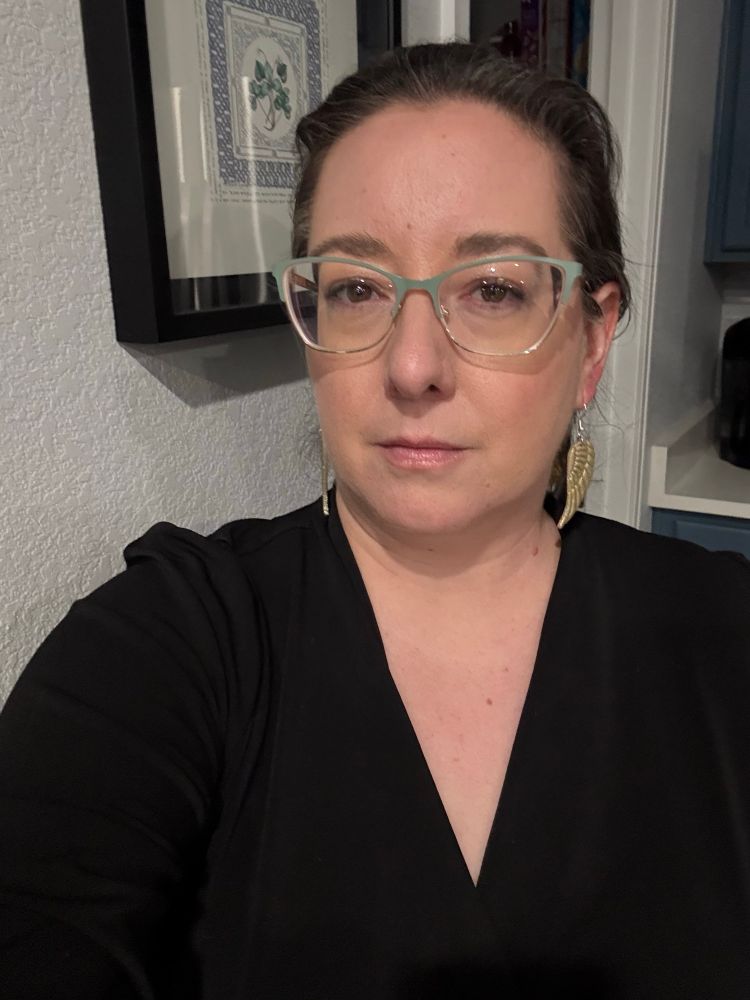Me in a black surplice top, hair braided back, glasses, gold wing earrings