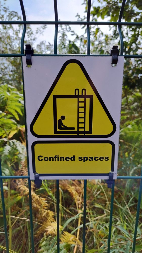 A photo of the "Confined Spaces" warning triangle. It is a yellow triangle with a black box on it with a ladder going inside. There is a very sad looking silhouette slumped against one wall.
