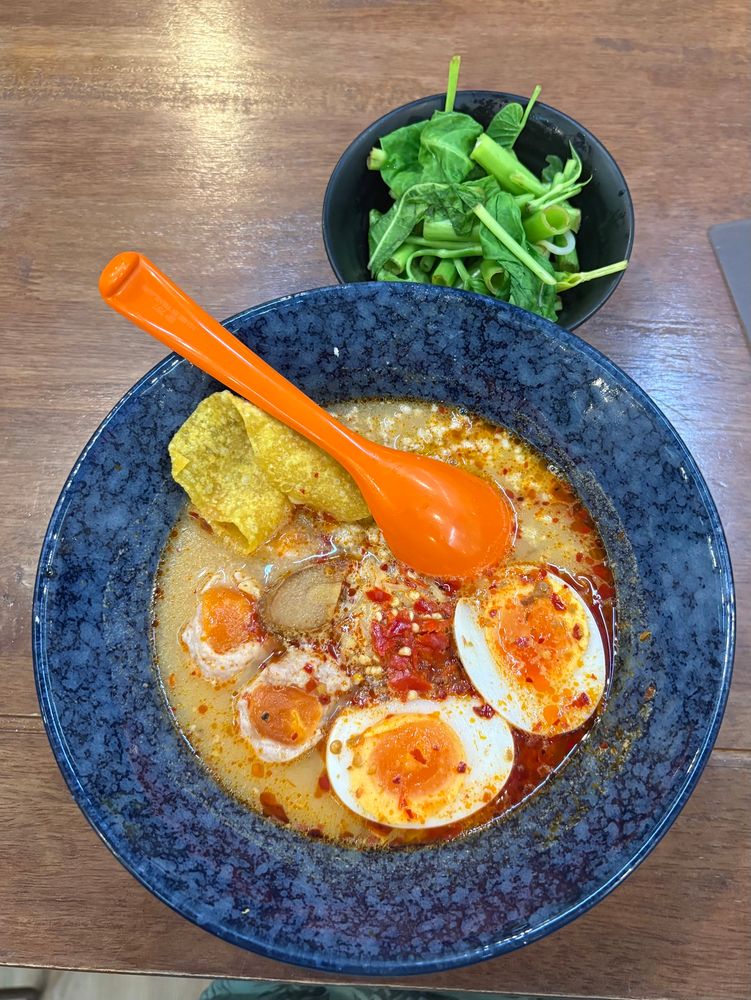 A blue bowl of Creamy Tom Yum Soup nooldes with Salted Egg at Piranya @ Siam, Bangkok. On the side there’s a serving of Morning Glory. 