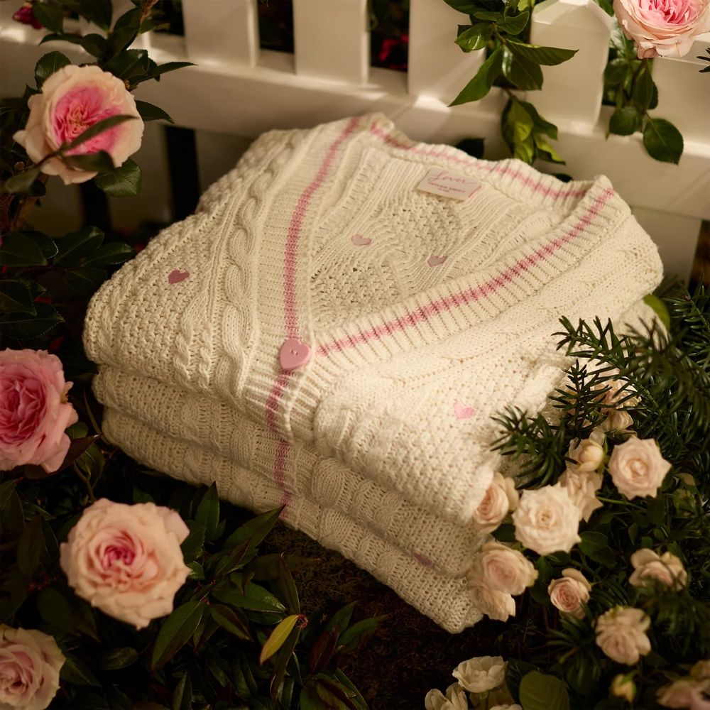three lover cardigans folded and stacked near a white picket fence with lots of roses surrounding them