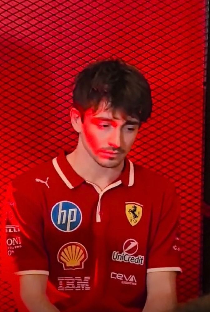 a photo of charles leclerc in a red 2025 team kit shirt in a red background. There’s a red light shining on one half of his face and he looks like the most depressed little mouse to have ever lived