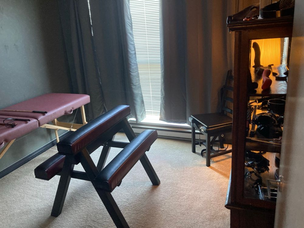 A small room with black curtains. Along The left wall is a massage table covered with floggers paddles and whips. In the Centre is a black spanking bench
