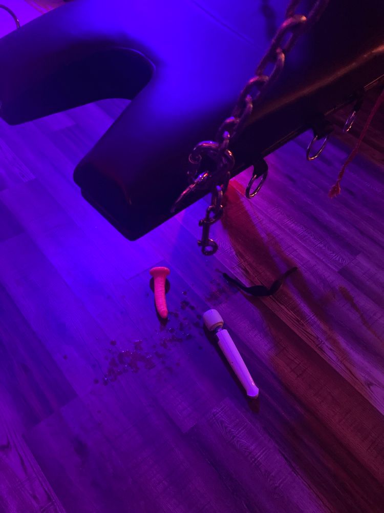 Pictured from above. In the foreground is a glimpse of a leather sex sling. On the floor beneath it is a splattered puddle, and laying beside the puddle is a hitachi magic wand and a pink dildo. 