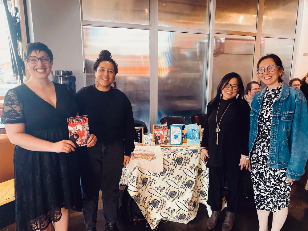 Four smiling people stand in front of a merchandise table with books on it. Sara Campos-Silvius is on the far left. She wears a black dress and holds up a copy of the zine containing her poetry. The zine cover is red and reads Hungry Zine. 