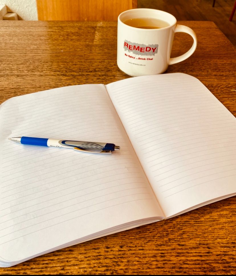 A notebook, pen, and white mug rest on a brown wooden cafe table. The notebook is open to a blank page. 