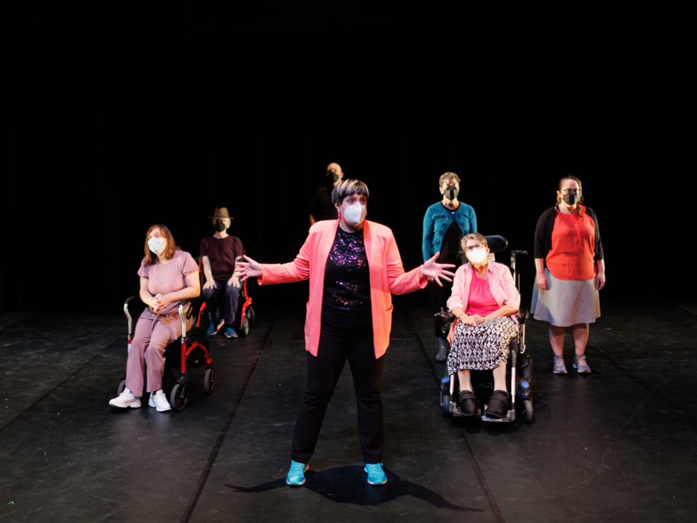 Sara Campos-Silvius stands facing forward in the centre and foreground of a brightly illuminated black box theatre stage. She wears a bright pink blazer and her hands are stretched wide as she engages the audience. Behind her are six dancers in seated and standing ready positions. Some dancers use mobility tools such as walkers and wheelchairs. All dancers wear clothing in various shades of pink, blue, and black, as well as face masks. 