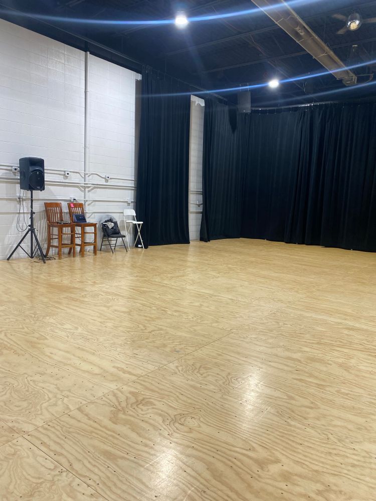 A brightly lit dance studio with wood coloured flooring, white walls, and a black curtain. Chairs and a music speaker line one of the walls. 