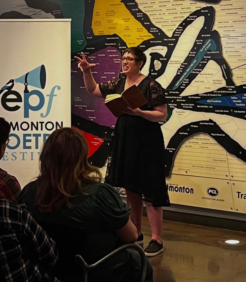 Sara Campos-Silvius stands in front of an audience performing poetry in a dramatic manner. She wears a black dress and holds a leather bound book. A banner to her side portrays the words: Edmonton Poetry Festival. 