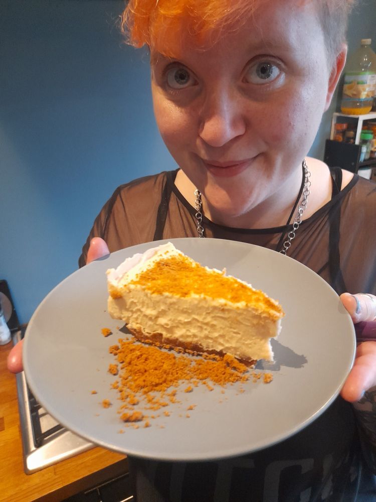 Camille is a white nonbinary person with blue eyes and pale pink short hair, wearing a mesh top, black dress, and silver chain necklace. They are holding a small grey plate with a slice of cheesecake on, which has biscuit crumbs sprinkled on top of it.