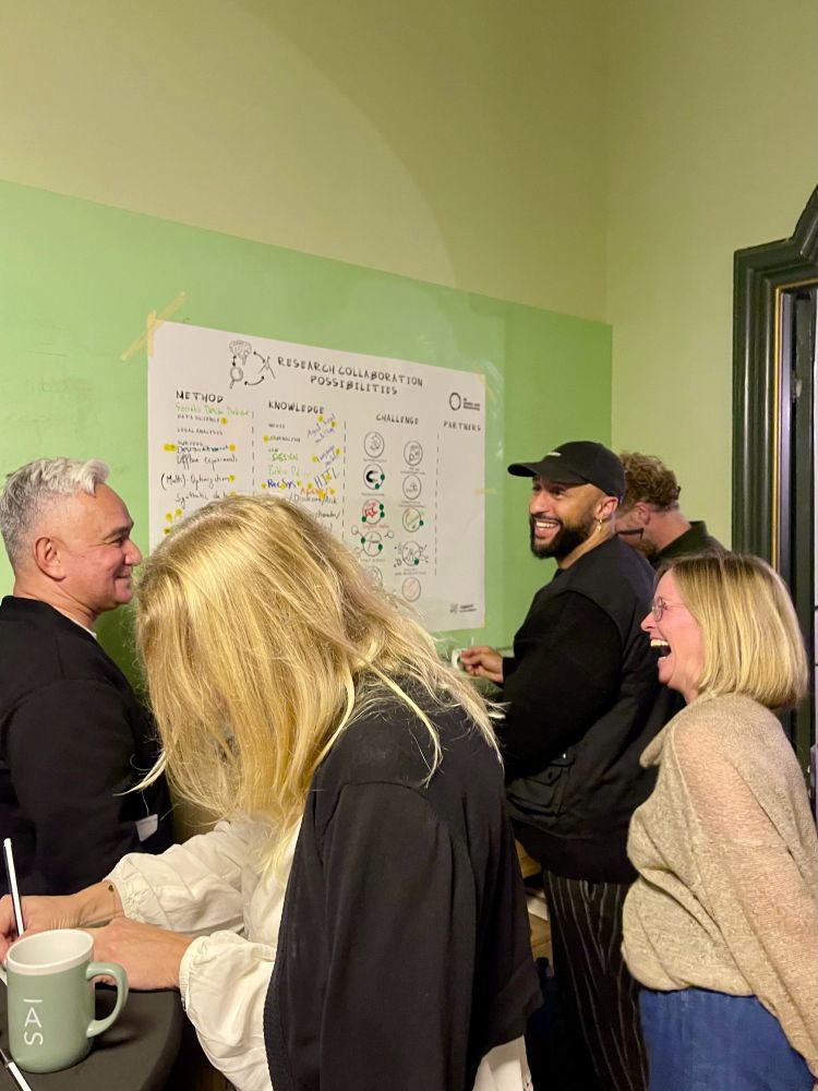Three partners are in conversation as they are standing in front of a poster. They are laughing and engaging with each other. One researcher is writing on a piece of paper in the front of the image. 
