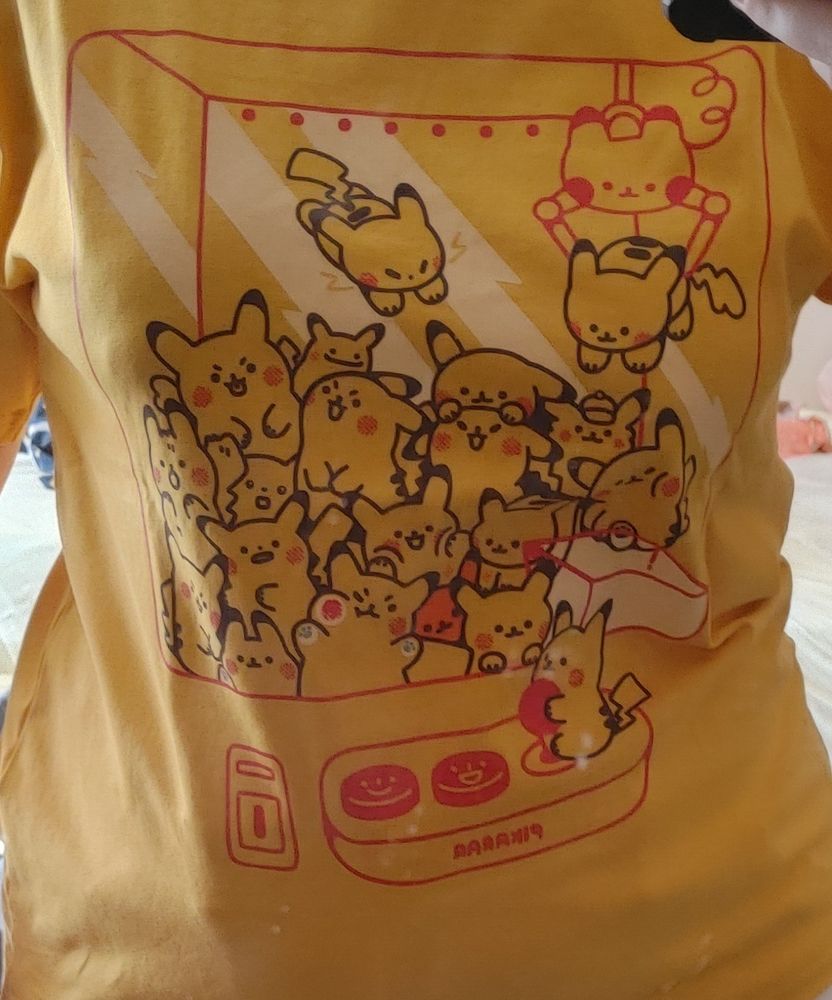 A strong yellow t shirt with a crane game filled with small pikachus. One of the pikachus is wearing a detective hat, and another is a ditto imposter. Theres a pikachu on the crane controller picking up another pikachu. One is squishing its cheek against the screen. T shirt was a preorder from pikarar on x.