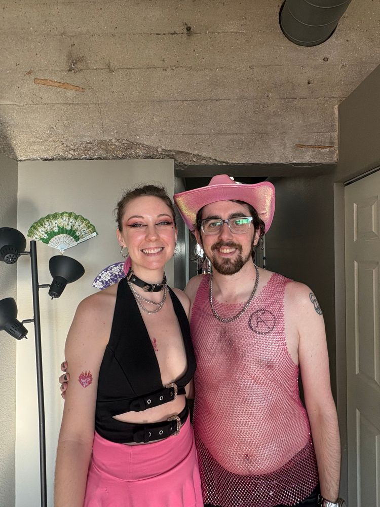 me & my partner before chappell roan’s concert. she’s wearing a black top, a pink skirt, and a choker necklace, with a pink “hot to go” temporary tattoo on her arm. i’m wearing a pink mesh tank top, black shorts, a pink cowboy hat, and a silver chain necklace. we are serving 