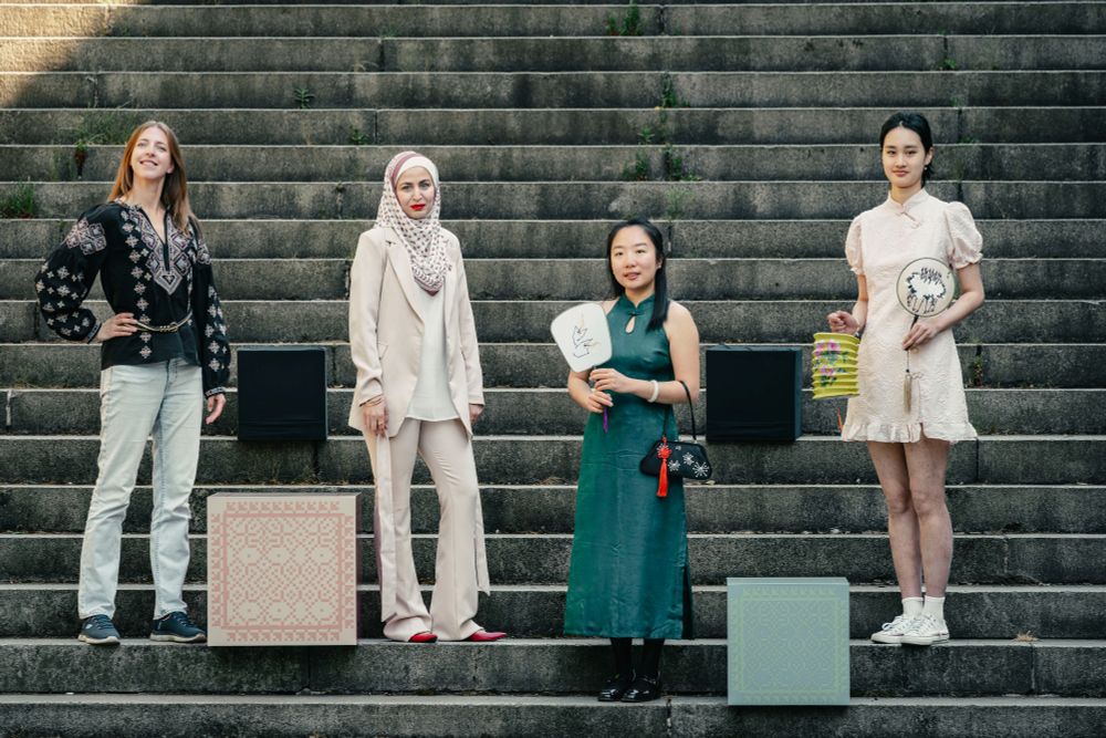 A diverse group of four smiling women stand on the steps at Kelvingrove Park. They are each wearing different types of traditional dress. Patterned boxes are stacked around them