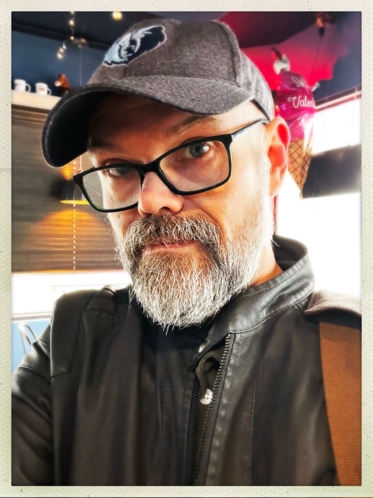 A man in a gray jacket, wearing a gray Memphis Grizzlies cap, standing in the queue at a coffee shop.