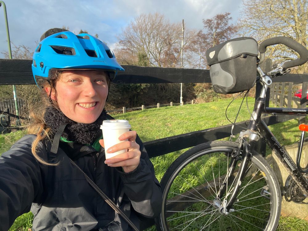 Veronica is sitting on the ground near her dad's bike. She is wearing her blue helmet and drinking a hot chocolate. 