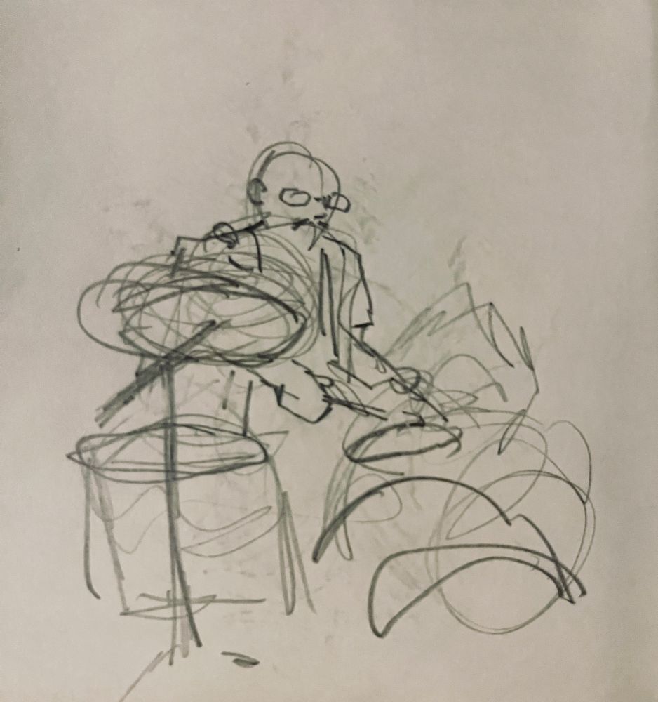 scribbly sketch of a drummer with a bald head, glasses, and a moustache. The drawing has a lot of simple circular shapes representing the drums and very simple features 