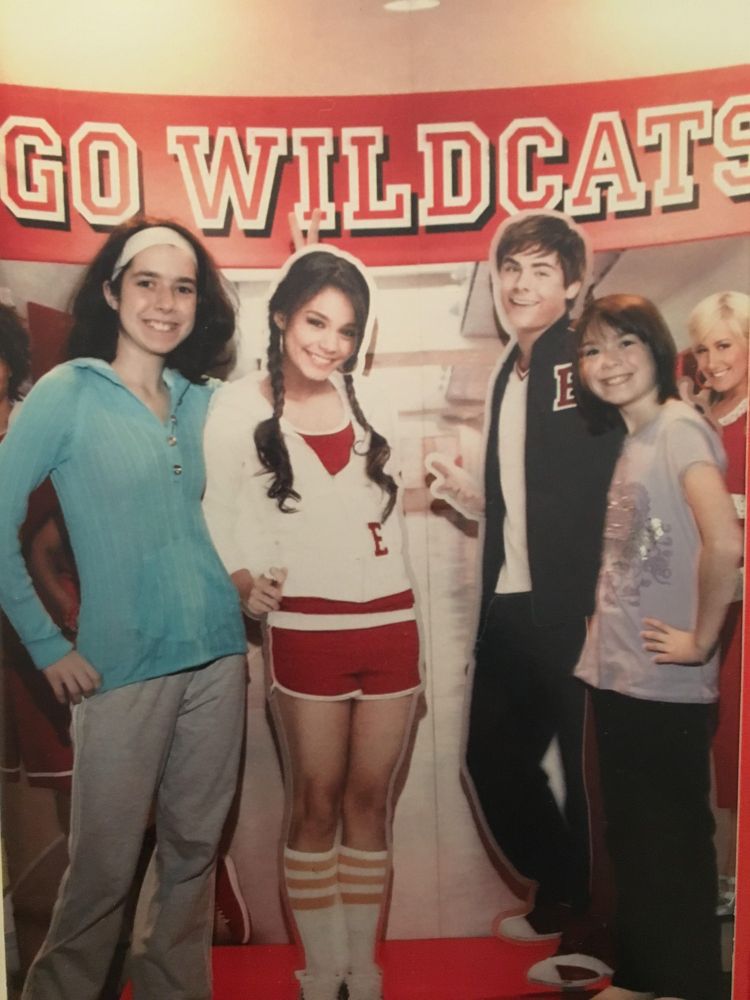 Cartboard cutouts of Gabriella Montez (Vanessa Ann Hudgens) and Troy Bolton (Zac Efron) dressed in Wildcats high school gear in front of a banner that reads "GO WILDCATS." Standing to the left of the Gabriella cutout is a pale white girl with a white headband in their dark brown frizzy hair and an icy blue sweater and gray sweatpants. To the right of the Troy cutout is her younger sister, a white girl with brown hair in a bangs and bob, donning a light purple T-shirt and black sweatpants.