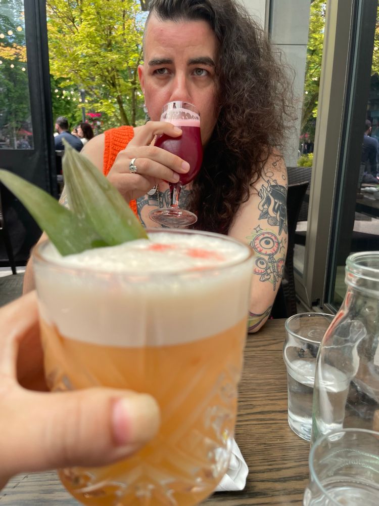 A picture of the same outdoor patio in this time M.L is drinking a sour beer. There is a hand holding some kind of cocktail in the foreground 