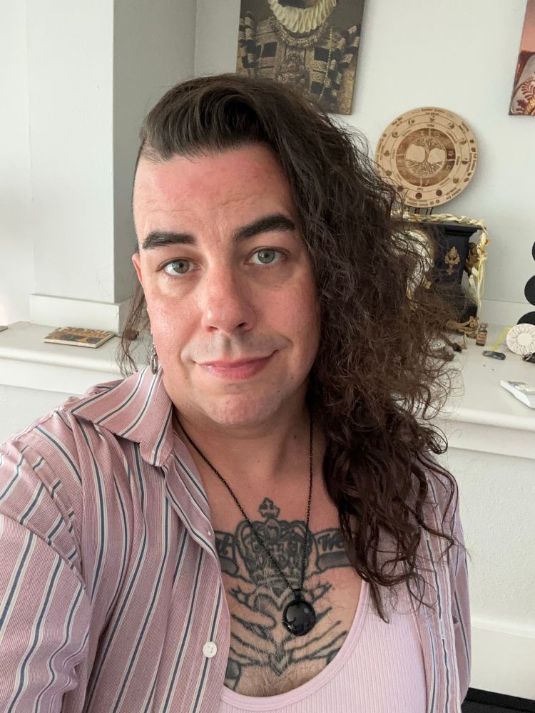 A selfie of ML. She’s wearing a pink tank and a pink, blue and white vertically striped button down. They are looking into the camera