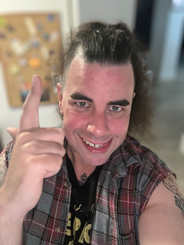 A selfie of ML. She is wearing a black t shirt with gold lettering and a red, grey, and black plaid flannel. Both have their sleeves cut off. She is making a mischievous face while making finger guns