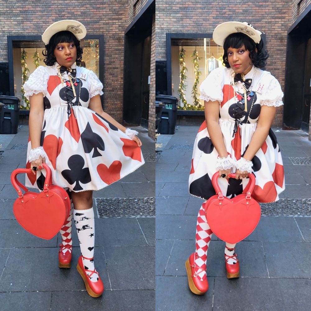 Milkcircus wearing a white lolita dress with red and black card suits all over it. They're wearing a white short sleeve blouse, white wristcuffs, bonnet with gold screenprinted crown, mismatched socks (red diamond & white with black card suits), and red wood platform shoes. They're holding a red bag shaped like a heart.