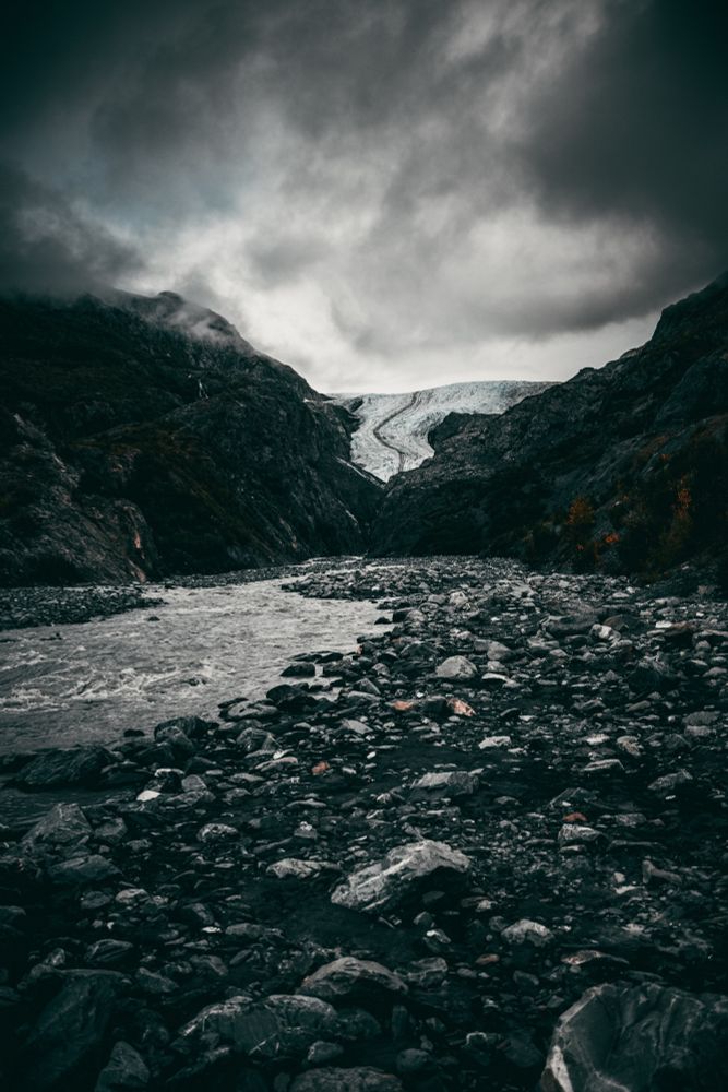 Primarily grayscale landscape photo of Exit glacier. The ground is rocky with a river flowing on the left side of the photo. The photo is taken in the river valley, looking up the valley towards the glacier. 