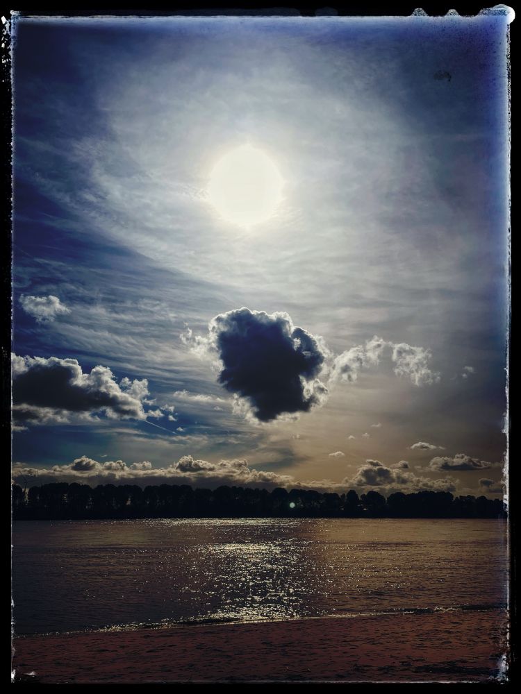 Foto von einem Blick auf den Fluss, darüber Wolken und die Sonne.