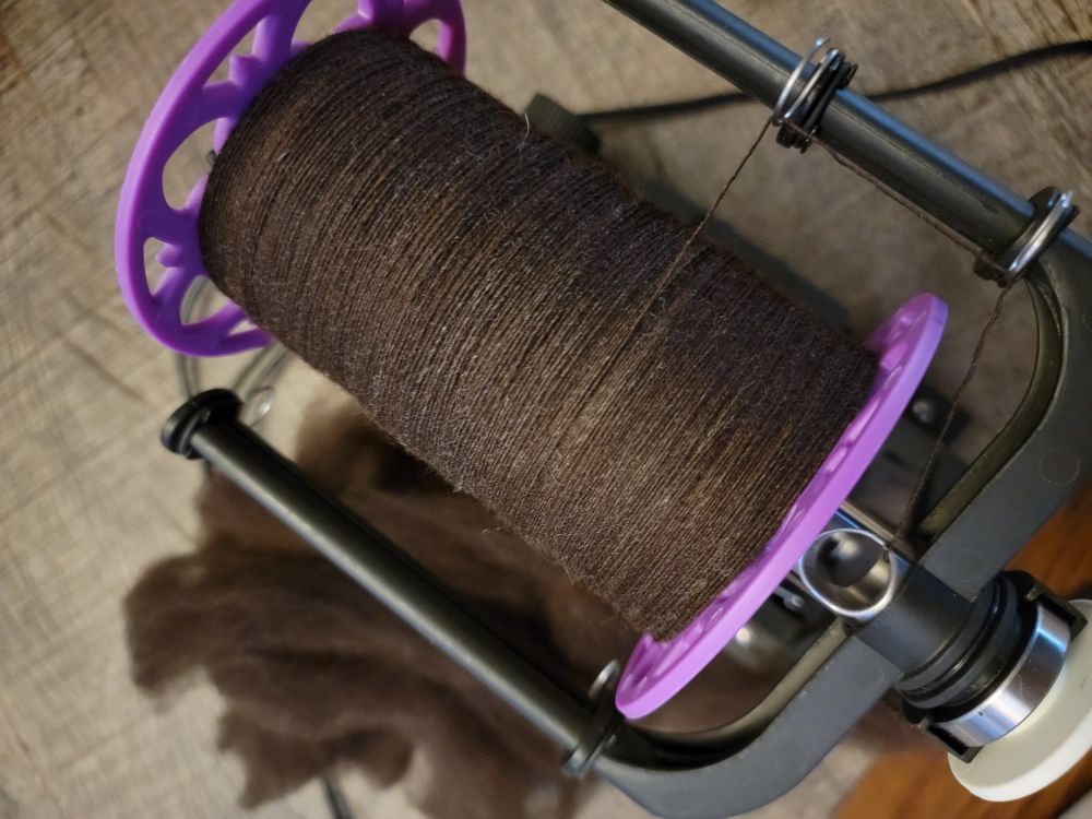 A purple bobbin sits on an EEW6 e-spinner partially filled with rich chocolate brown Merino x Finn wool. A portion of hand-combed spun fiber that has yet to be spun up rests on the table below the flyer slightly out of focus.