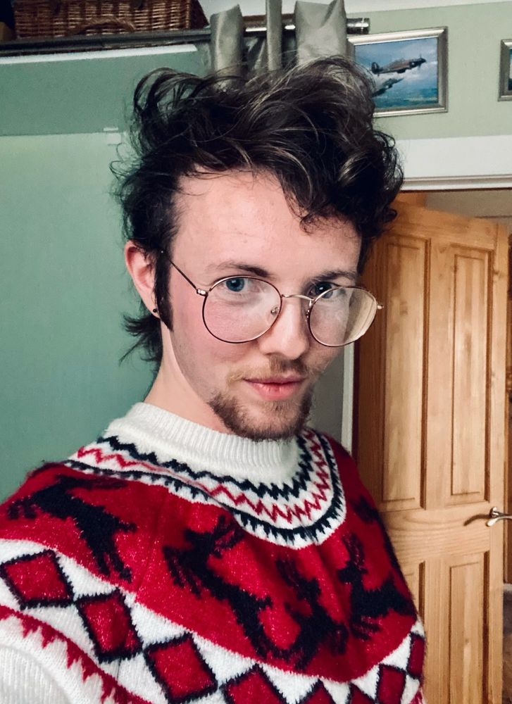 Me as I look now - with longer curly hair, goatee beard and moustache, and long sideburns. I’m wearing a Christmas jumper, white with black and red diamond and zigzag patterns, and reindeer silhouettes across the chest and arms. My chest is unbound - some slight breast curvature is proudly visible. 