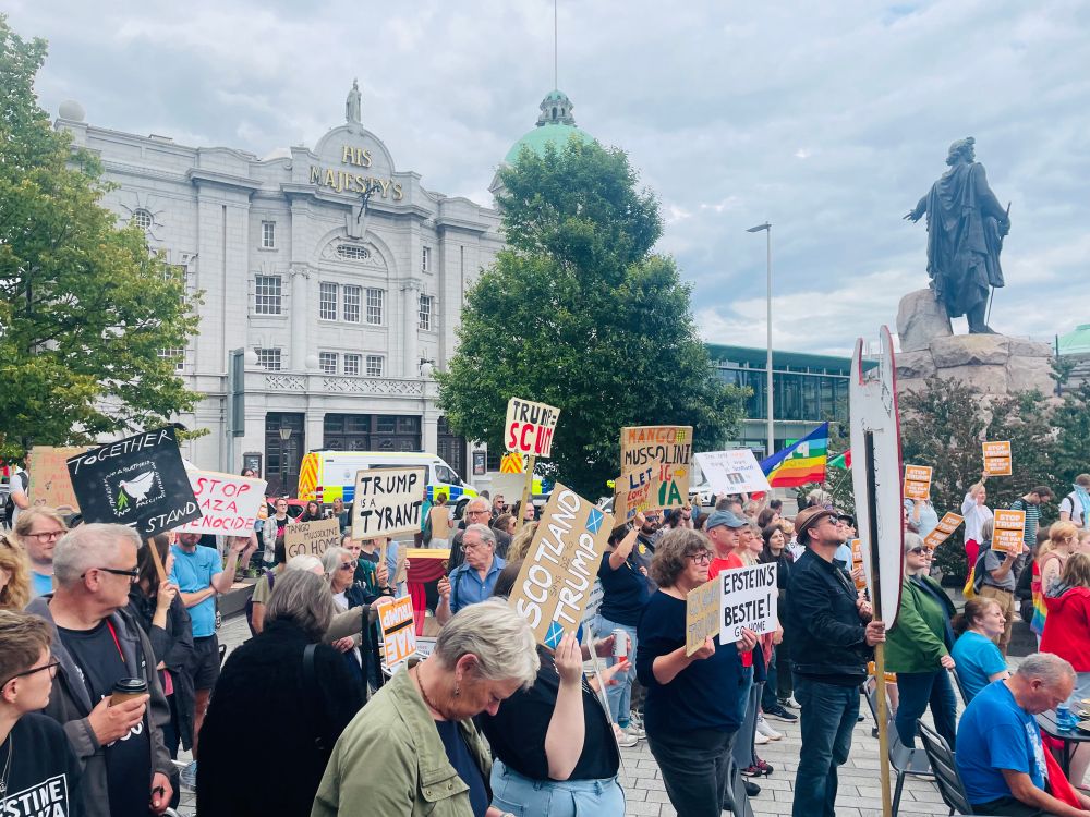 A crowd of people can be seen holding signs that say - “Trump Tyrant”, “Epstein’s Bestie”, “Trump = Scum”, and “Together We Stand”