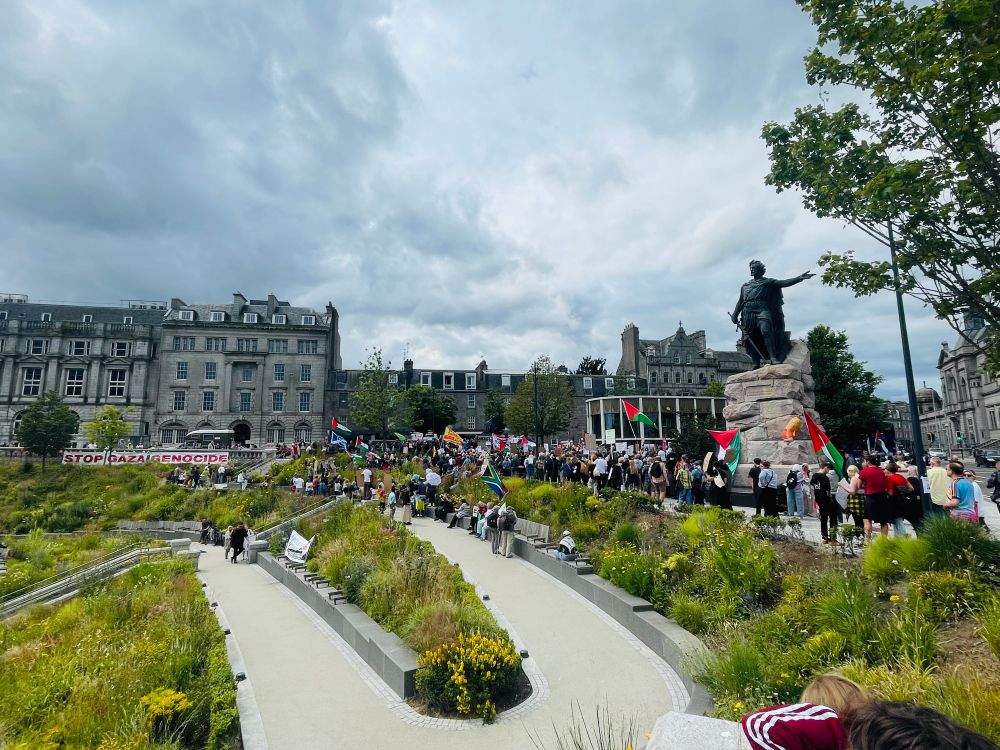 The crowd on Union Terrace Gardens can be seen and a large “Stop Gaza Genocide” banner hangs in the background.