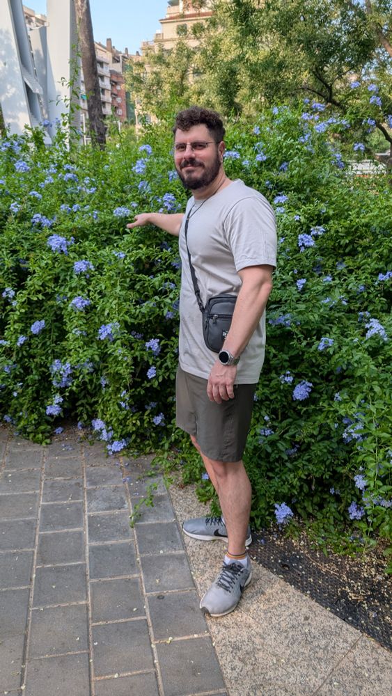 Eu pousando para a foto ao lado de um arbusto com flores azuis imitando uma pose comum de senhores da terceira idade.