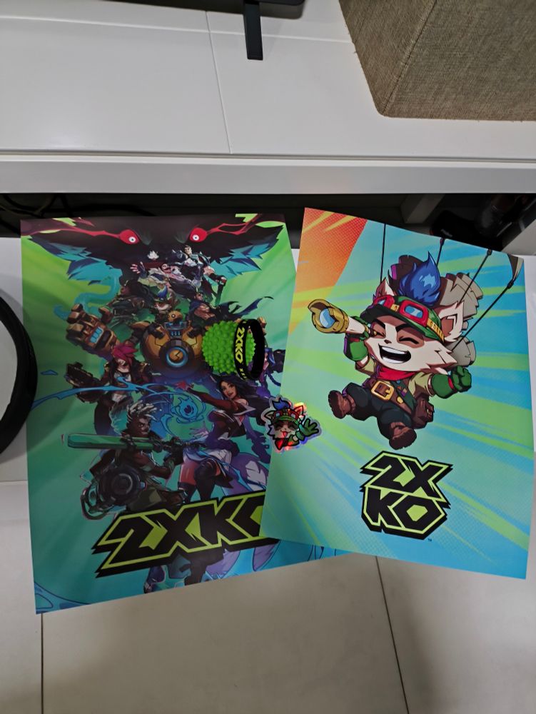 2 cartazes de 2xko, um com todo o elenco e um com Teemo (criatura pequena e felpuda com roupa de escoteiro), um adesivo brilhante também de Teemo, uma pulseira de borracha preta e uma bola de estresse/cachorro verde e com "espinhos" grossos, como um maxixe.