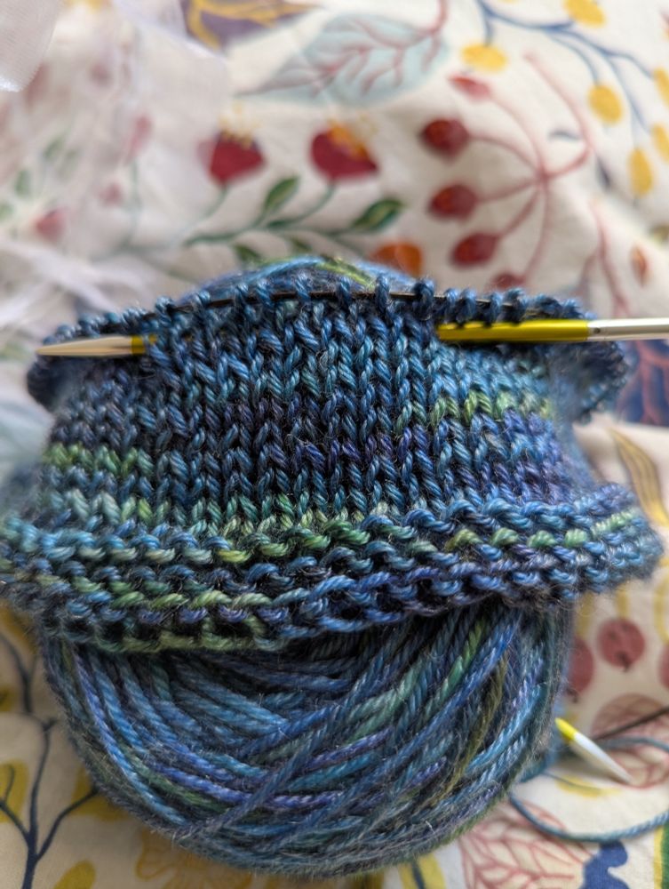 Blue green variegated tencel/merino yarn with a few rows of garter and the more rows of stocking stitch, skewered to its yarn cake with a yellowish knitpro needle