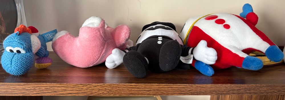 A handful of plushie objects all laying on the backs like they got wiped out. A Blue Yoshi Amiibo. A pink teddy bear. A Jack Frost and Teddy plushie from Persona/SMT 
