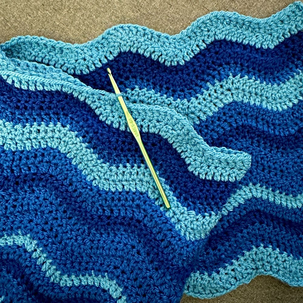 A work-in-progrss crochet blanket with wavy blue stripes and a green aluminum crochet hook.