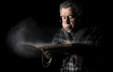 vieil homme tenant un vieux livre dans ses mains soufflant fort dessus ce qui a pour effet de le dépoussiérer